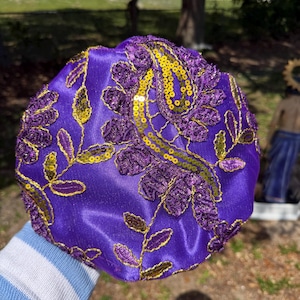 May include: A round, purple satin bonnet with intricate embroidery and embellishments. The design features floral patterns and gold sequins. The bonnet is a vibrant shade of purple, with gold and purple accents.