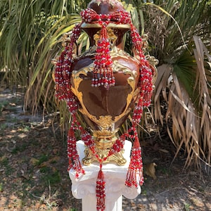 May include: A large, ornate brown and gold vase with a lid, adorned with intricate gold detailing. Strands of red beaded tassels cascade down the sides. The vase is displayed on a white pedestal, set against a backdrop of lush greenery.