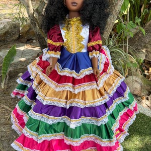 May include: A doll with dark skin and black curly hair, wearing a colorful layered dress. The dress features red, gold, purple, green, and pink tiers, trimmed with white lace and gold accents. The doll has a gold headband and a detailed bodice.