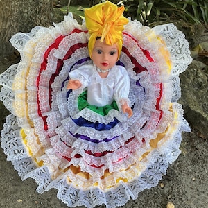May include: A doll wearing a traditional, colorful dress with layers of red, yellow, blue, and green. The dress is trimmed with white lace. The doll has a yellow head wrap and a white blouse.