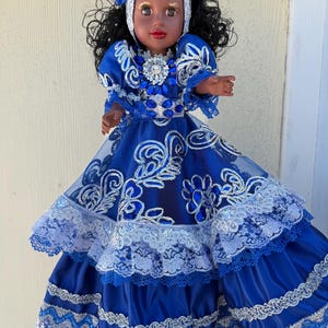 May include: A doll wearing a vibrant blue dress with silver and white accents. The dress features intricate embroidery, lace trim, and layered ruffles. The doll has dark hair, a white head covering, and a floral crown. A large jewel adorns the neckline.