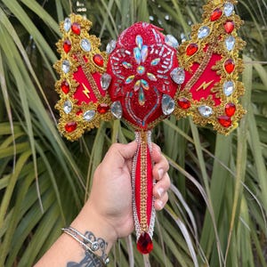 May include: A red and gold decorative scepter with ornate detailing. The scepter features a central red jewel, surrounded by gold trim, red and clear gemstones, and a lightning bolt design. The handle is embellished with red fabric and clear beads.
