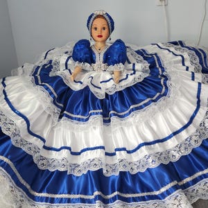 May include: A doll dressed in a traditional blue and white satin dress with lace trim. The doll has a matching blue head covering and a detailed bodice. The dress features layers of fabric and silver accents, creating a formal appearance.