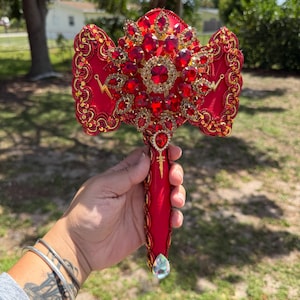 May include: A red and gold decorative scepter with a large, ornate head adorned with red gemstones and gold accents. The handle is red with gold trim and a teardrop-shaped gem at the base. The scepter is held in a hand.