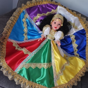 May include: A doll with black hair and a gold headband, wearing a colorful dress. The dress is a patchwork of red, green, yellow, blue, purple, and white satin, trimmed with gold lace. The doll's outfit includes a white blouse with gold accents.