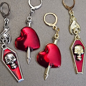 May include: Four pairs of earrings with a gothic theme. Each pair features a red heart with a silver or gold dagger piercing through it, and a coffin with a skull design. The earrings are silver or gold toned.