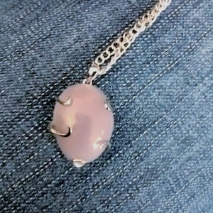 May include: A silver chain necklace with a light pink oval pendant. The pendant is set in a silver frame with a decorative design.