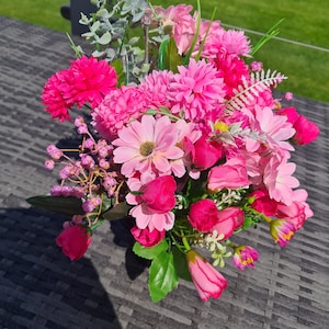 May include: A colourful artificial flower arrangement featuring pink and fuchsia blooms. The bouquet includes various flower types, such as daisies, roses, and chrysanthemums, with green foliage. The arrangement is set on a dark surface.