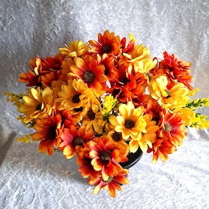 May include: A dense bouquet of artificial flowers featuring vibrant shades of yellow, orange, and deep red petals. The blossoms have dark brown centres and are arranged in a rounded cluster, with small yellow sprigs interspersed throughout the stems.