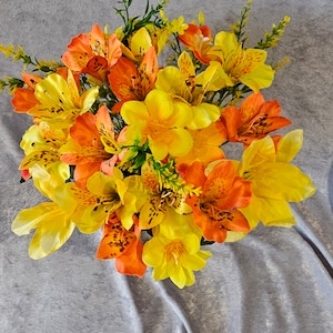 May include: A bouquet of yellow and orange artificial flowers with brown spots on the petals. The flowers are arranged in a circular shape and are set against a grey background.