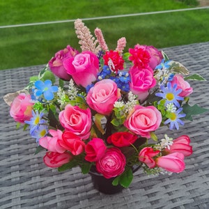 May include: A bouquet of pink, red and blue artificial flowers in a black pot. The flowers are arranged in a circular shape and are surrounded by green leaves.