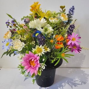 May include: A colourful artificial flower arrangement, including daisies, lilies, and other blooms in yellow, white, orange, pink, and purple. The flowers are arranged in a black top hat, creating a unique and eye-catching display. A decorative floral centrepiece.