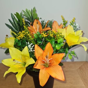 May include: A bouquet of artificial flowers in a black vase. The bouquet includes orange and yellow lilies with green foliage.