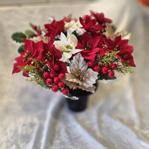 May include: A festive floral arrangement featuring vibrant red poinsettias, white flowers, and faux red berries. The arrangement includes silver and gold-toned leaves and green sprigs, all set in a black vase. A seasonal holiday decoration.