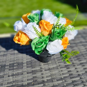 May include: A bouquet of artificial flowers in a black pot. The flowers are white, orange, and green, and they are arranged in a circular pattern.