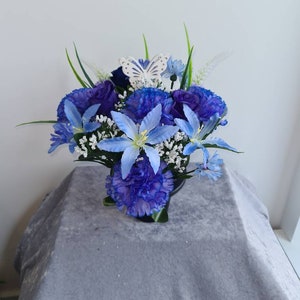 May include: A blue and white artificial flower arrangement with a white butterfly decoration. The arrangement is in a black vase and is sitting on a grey surface.
