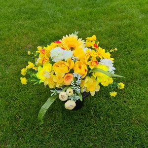 May include: A bouquet of yellow and white flowers in a black vase. The flowers are arranged in a circular shape and are surrounded by green leaves.