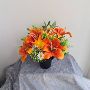 May include: A colourful floral display of orange lilies with yellow centres, complemented by green leaves and small white flowers. The arrangement is presented in a black pot, offering a bold contrast.