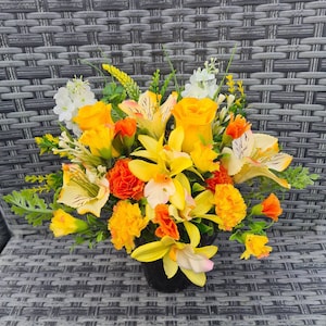May include: A yellow and orange artificial flower arrangement in a black pot. The flowers include roses, lilies, and carnations.