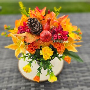 May include: A colourful autumn floral display with orange lilies, marigolds, and red dahlias. A pinecone and speckled red gourds are among the flowers. The arrangement is in a black metal container on a wooden slice.
