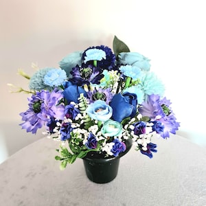May include: A blue and purple artificial flower arrangement in a black pot. The flowers are made of silk and are arranged in a bouquet.