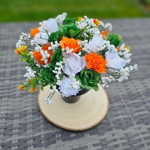 May include: A bouquet of white and orange flowers arranged in a black vase. The flowers are surrounded by small white flowers and green leaves.