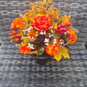 May include: A colourful floral display in a black vase, showcasing orange, red, and yellow flowers, berries, and leaves. The arrangement includes a large, multi-petaled flower, with accents of white and burgundy, perfect for autumn.