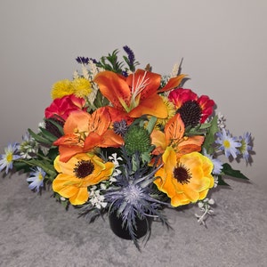 May include: A colourful artificial flower arrangement in a black pot. The arrangement features orange lilies, yellow and orange poppy-like flowers, red flowers, and blue thistle-like flowers.
