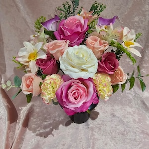 May include: A vibrant floral display with artificial roses, carnations, and other flowers in pink, white, and peach hues. The arrangement is set in a dark vase, with green foliage. The flowers are arranged to create a full, textured look.
