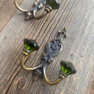 May include: Two decorative wall hooks with a vintage aesthetic. Each hook features a curved brass arm and a faceted, emerald-green glass knob. The ornate backplates have a dark, antique finish, adding to the vintage charm. The hooks are mounted on a weathered wooden surface.