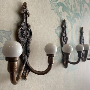 May include: Ornate bronze wall hooks with white ceramic knobs. The hooks have a decorative, vintage design and are mounted on a patterned wall. The hooks are ideal for hanging coats, hats, or other items.