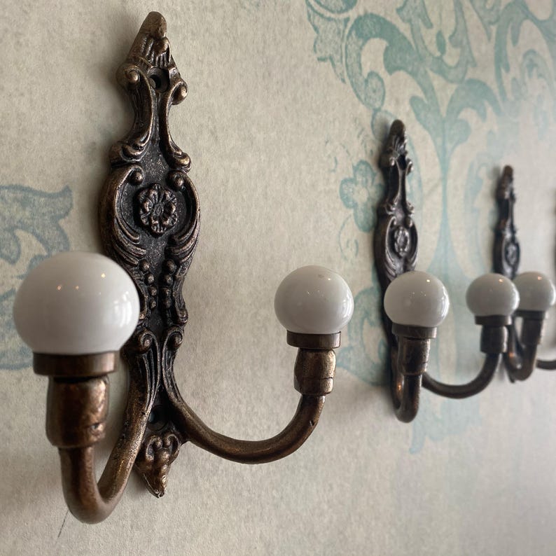 May include: Ornate bronze wall hooks with white ceramic knobs. The hooks have a decorative, vintage design and are mounted on a patterned wall. The hooks are ideal for hanging coats, hats, or other items.