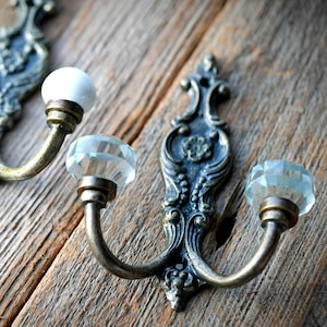 May include: Ornate, antique-style wall hooks. The hooks have a bronze finish with decorative backplates and curved arms. One hook features a white ceramic knob, while the others have faceted, clear glass knobs. The hooks are mounted on a weathered wooden surface.