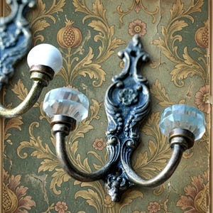 May include: Ornate, antique-style wall hooks with a distressed finish. The hooks feature decorative backplates and curved arms, topped with clear, faceted crystal knobs and a white knob. The hooks are mounted on a vintage floral wallpaper.