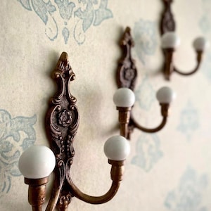 May include: Three wall-mounted hooks featuring a bronze-toned metal base with ornate, scroll-like detailing and a central floral motif. Each hook has two upward-curving arms, each tipped with a smooth, round, white ceramic ball finial.