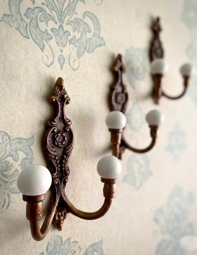 May include: Three wall-mounted hooks featuring a bronze-toned metal base with ornate, scroll-like detailing and a central floral motif. Each hook has two upward-curving arms, each tipped with a smooth, round, white ceramic ball finial.