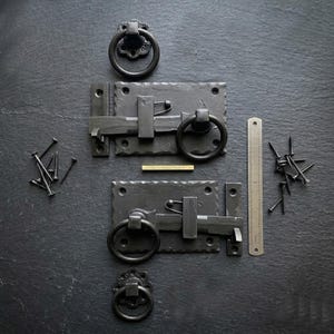 Könnte beinhalten: Schwarze schmiedeeiserne Türriegel mit Ringgriffen und passenden dekorativen Ringzügen. Inklusive Schrauben und Lineal zur Maßstabsbestimmung. Die Hardware wird auf einer dunkelgrauen Oberfläche präsentiert.