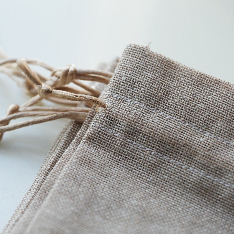 Op de afbeelding: Een close-up van een beige trekkoordtas gemaakt van natuurlijke stof met een geweven textuur. De tas is dubbelgevouwen en het trekkoord is bovenaan vastgeknoopt.