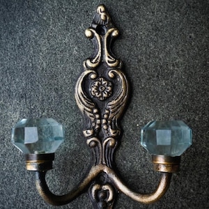 May include: An ornate, antique-style wall hook with a dark bronze finish and two curved arms. Each arm supports a faceted, light blue glass knob. The hook features decorative floral and scrollwork details, adding a vintage aesthetic.