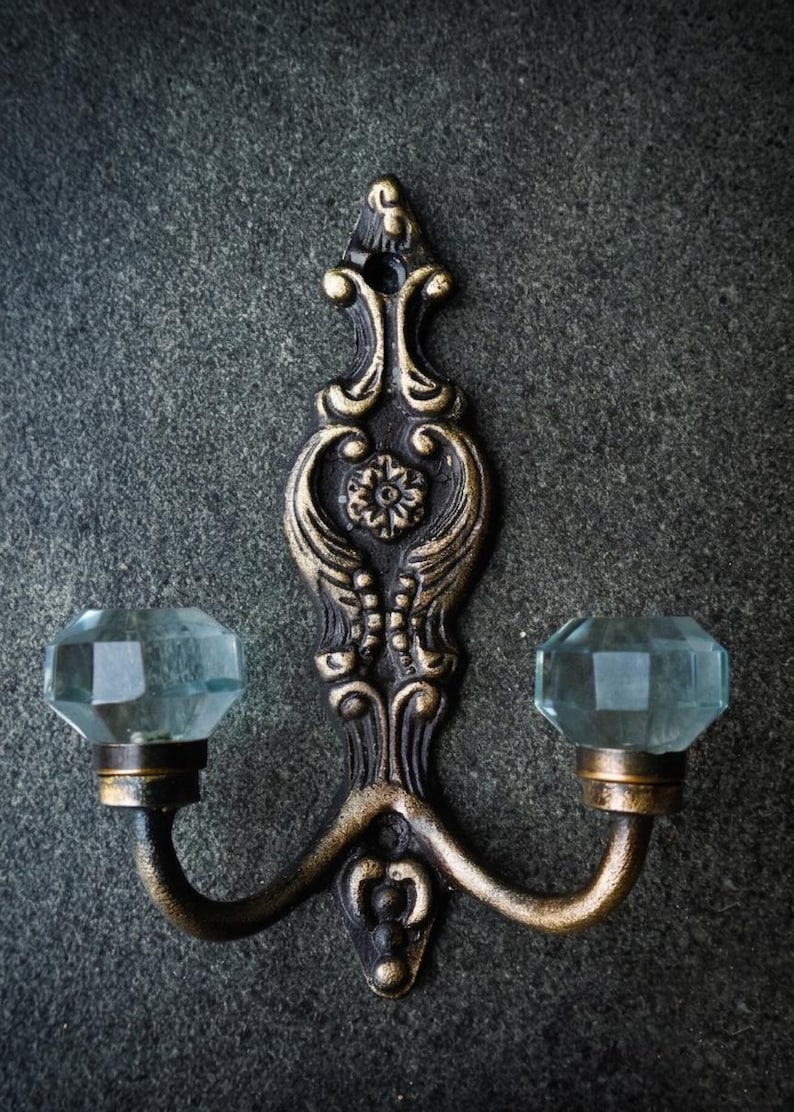 May include: An ornate, antique-style wall hook with a dark bronze finish and two curved arms. Each arm supports a faceted, light blue glass knob. The hook features decorative floral and scrollwork details, adding a vintage aesthetic.