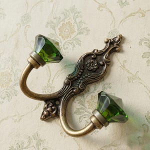 May include: An ornate, antique-style wall hook with two curved arms. Each arm features a faceted, emerald-green crystal. The hook's metalwork has a bronze finish with decorative floral and scroll details, adding a touch of vintage elegance.