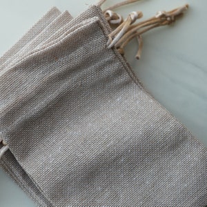 Op de afbeelding: Een set van vier kleine, natuurlijke kleur jute zakken met trekkoord. De zakken zijn op elkaar gestapeld en de trekkoorden zijn aan elkaar gebonden.