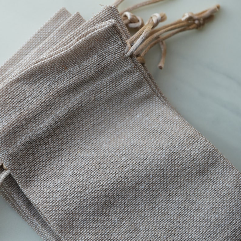 Op de afbeelding: Een set van vier kleine, natuurlijke kleur jute zakken met trekkoord. De zakken zijn op elkaar gestapeld en de trekkoorden zijn aan elkaar gebonden.