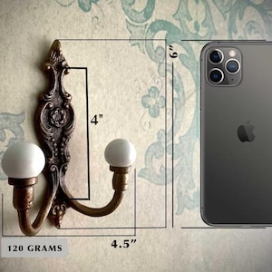 May include: Ornate bronze-colored wall hook with two white ball-shaped knobs. The hook is 4 inches tall and 4.5 inches wide. The weight is 120 grams. The hook is shown next to a phone for size comparison.
