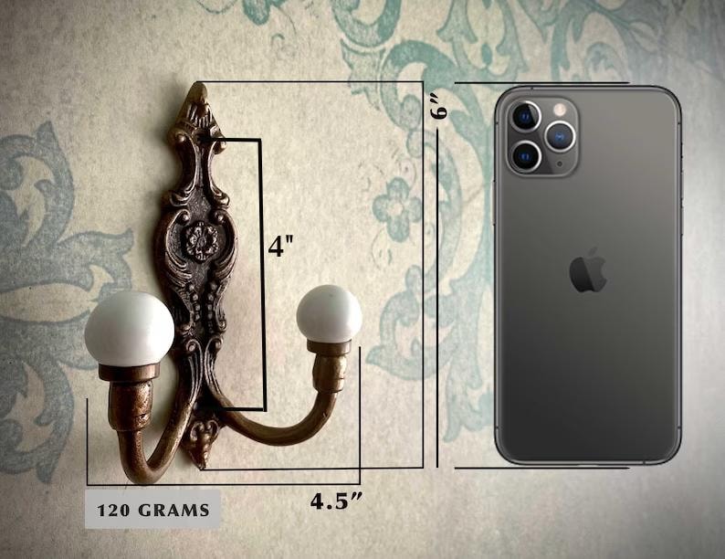 May include: Ornate bronze-colored wall hook with two white ball-shaped knobs. The hook is 4 inches tall and 4.5 inches wide. The weight is 120 grams. The hook is shown next to a phone for size comparison.