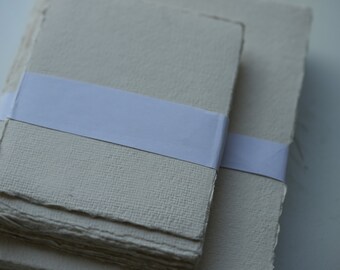 May include: Two stacks of handmade paper with rough, deckled edges, secured with a white paper band. The paper has a textured, off-white appearance, suggesting a natural, artisanal quality. Suitable for stationery or art projects.