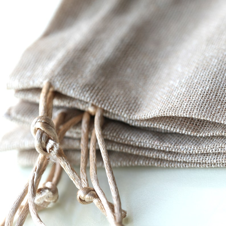 Op de afbeelding: Een stapel van vier kleine, beige, jute trekzakjes met bruine koorden. De zakjes zijn gevouwen en de koorden zijn geknoopt.