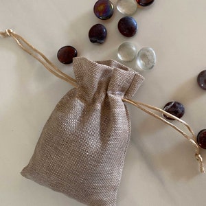 Op de afbeelding: Een klein, beige jute trekkoordtasje met een natuurlijk touwtje. Het tasje ligt op een wit oppervlak met verschillende kleine, ronde glazen steentjes die eromheen verspreid liggen. De steentjes zijn in tinten bruin, helder en iriserend.