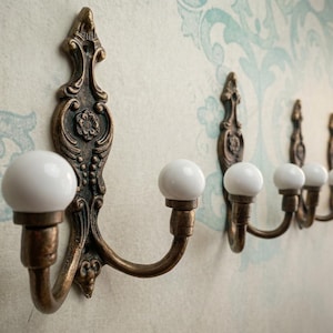 May include: A row of antique bronze wall hooks with white ceramic knobs. The hooks have ornate, decorative backplates and are mounted on a wall with a floral pattern. The hooks are ideal for hanging coats, hats, or other items.