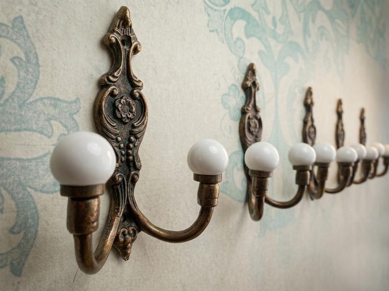 May include: A row of antique bronze wall hooks with white ceramic knobs. The hooks have ornate, decorative backplates and are mounted on a wall with a floral pattern. The hooks are ideal for hanging coats, hats, or other items.